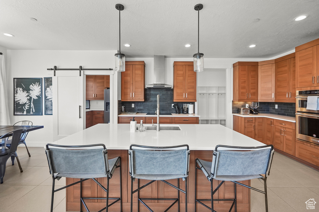 Kitchen featuring wall chimney range hood, brown cabinetry, stainless steel double oven, tasteful backsplash, and light countertops