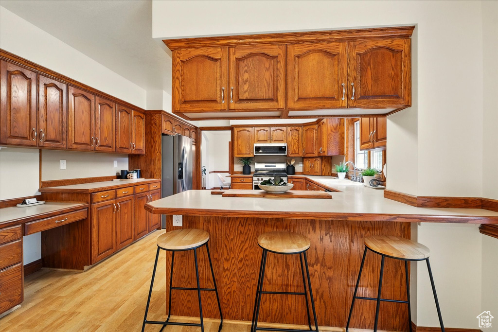 Kitchen featuring stainless steel appliances, light wood-style flooring, a peninsula, brown cabinets, and a kitchen bar
