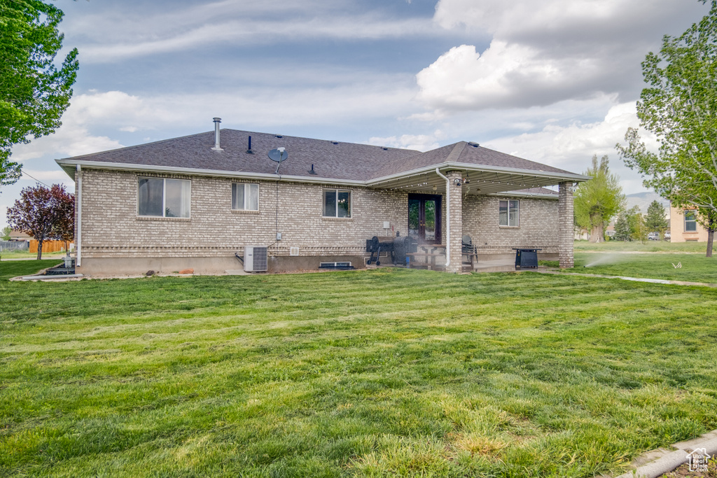 Back of property with central AC unit, brick siding, a lawn, roof with shingles, and a patio