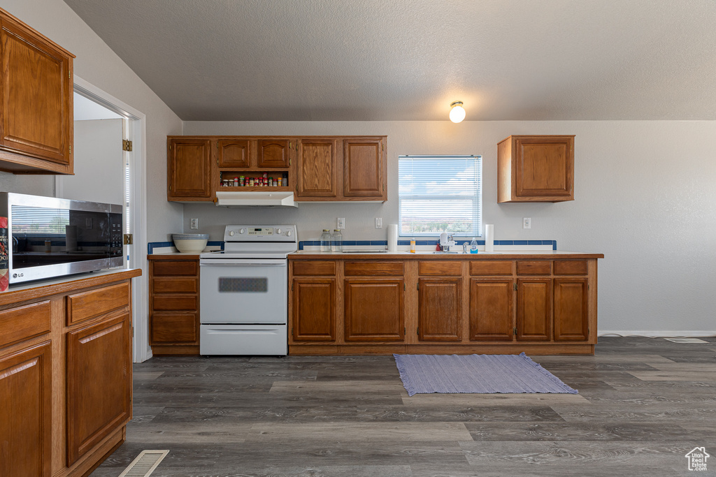 Kitchen with white electric range oven, stainless steel microwave, under cabinet range hood, brown cabinetry, and dark wood-type flooring