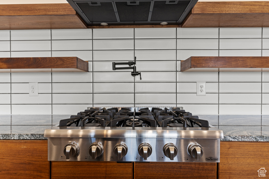 Kitchen view of stainless steel gas stovetop, ventilation hood, decorative backsplash, dark stone countertops, and pot filler
