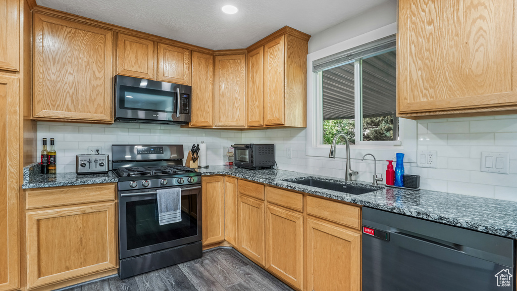 Kitchen with appliances with stainless steel finishes, a sink, backsplash, dark wood-style floors, and dark stone countertops