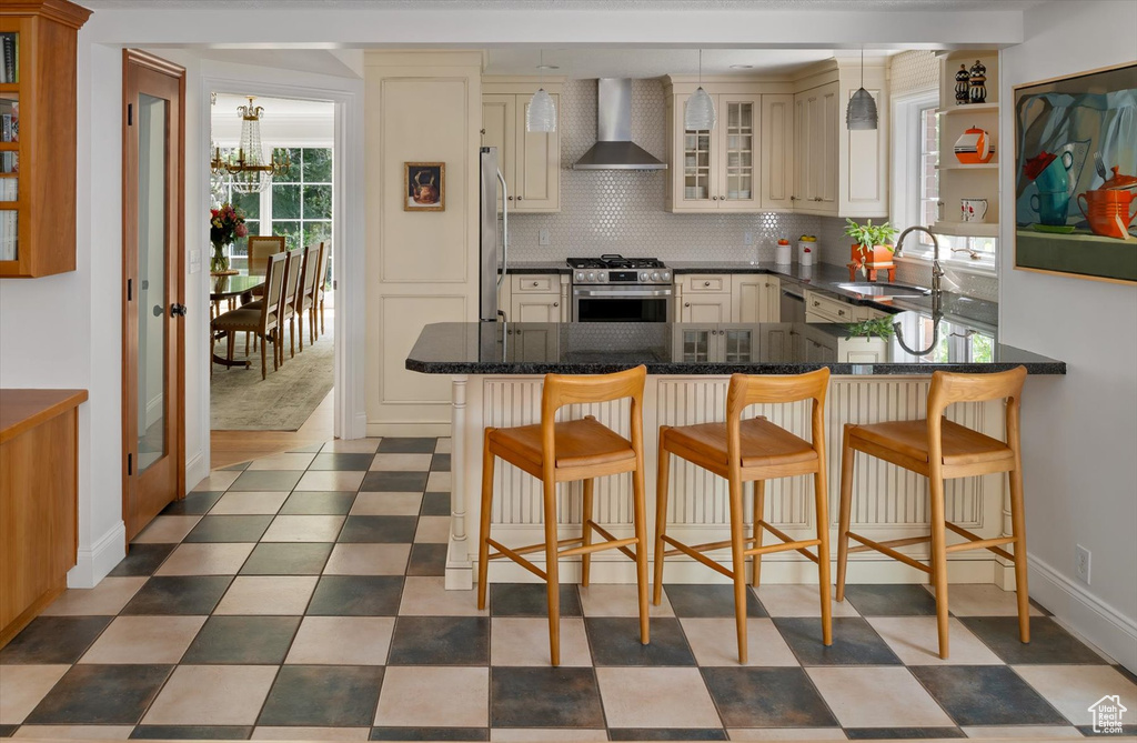 Kitchen with wall chimney exhaust hood, stainless steel appliances, cream cabinetry, a peninsula, and a sink