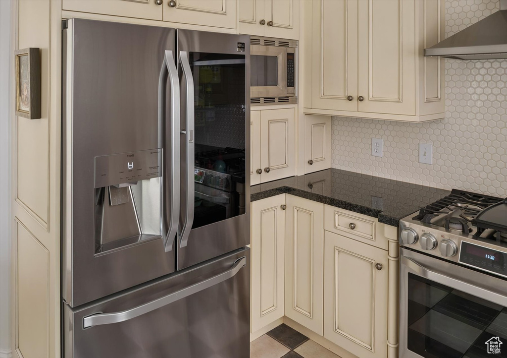 Kitchen featuring appliances with stainless steel finishes, cream cabinets, wall chimney exhaust hood, decorative backsplash, and dark stone counters