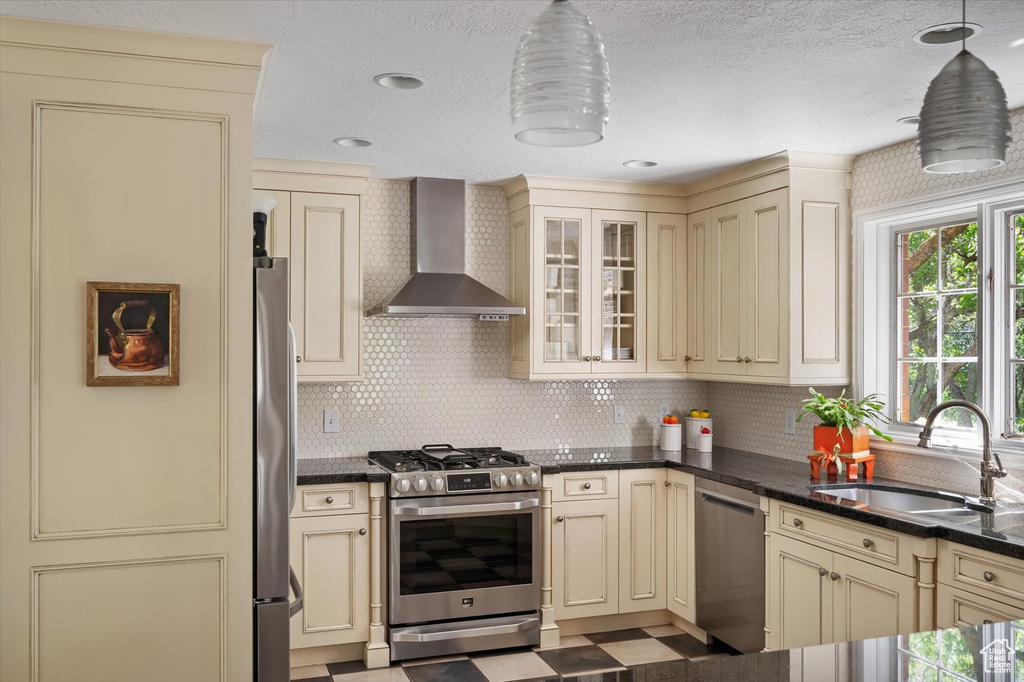 Kitchen with cream cabinetry, appliances with stainless steel finishes, wall chimney range hood, a sink, and decorative backsplash