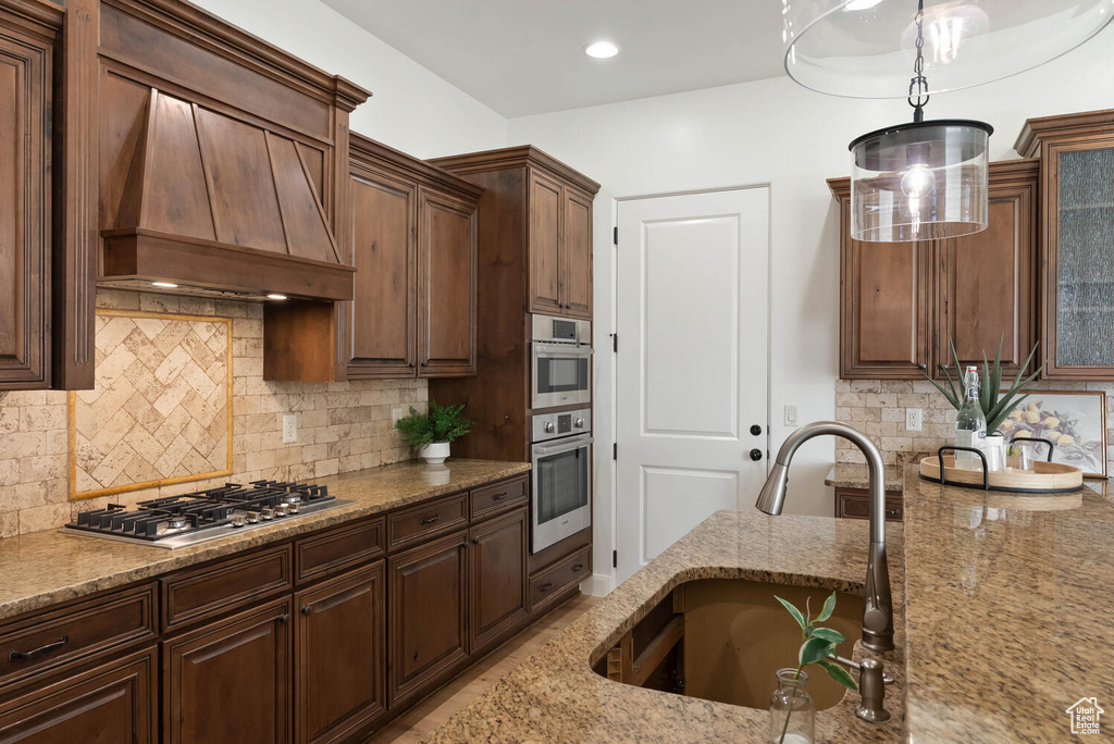 Kitchen with a sink, custom range hood, light stone counters, appliances with stainless steel finishes, and decorative backsplash