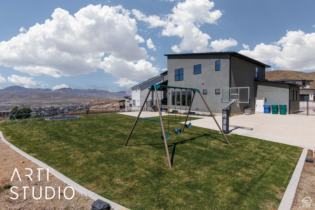 Back of house featuring fence, stucco siding, a mountain view, a playground, and a lawn