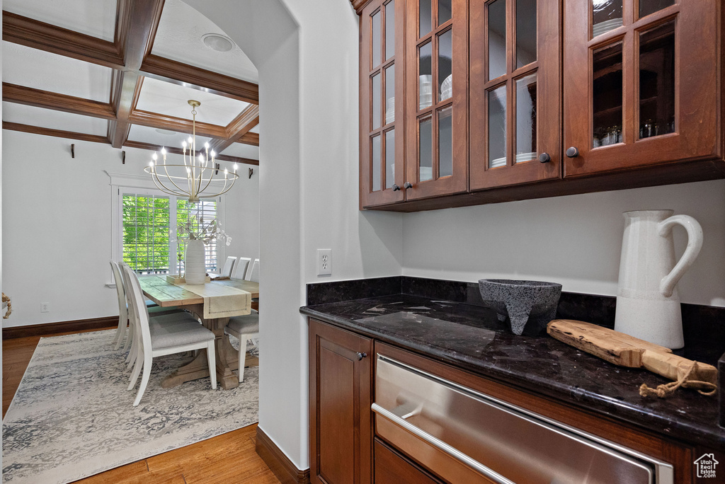 Kitchen with coffered ceiling, dark stone counters, wood finished floors, a chandelier, and glass insert cabinets