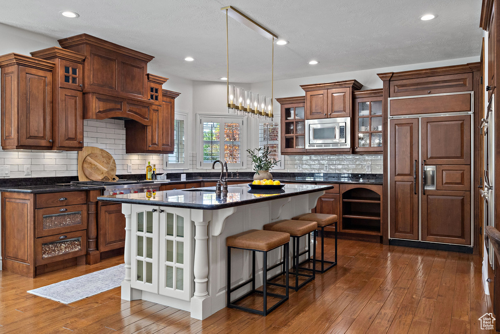 Kitchen featuring built in appliances, a breakfast bar area, a center island with sink, dark wood-style flooring, and tasteful backsplash