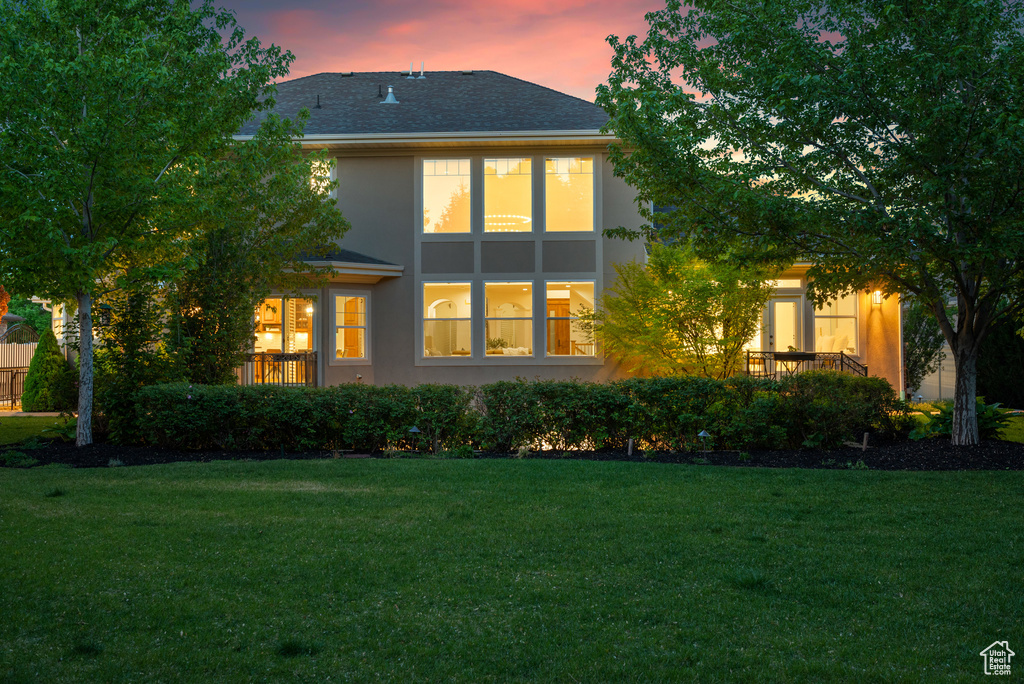 Back of house at dusk featuring stucco siding, a lawn, and a shingled roof
