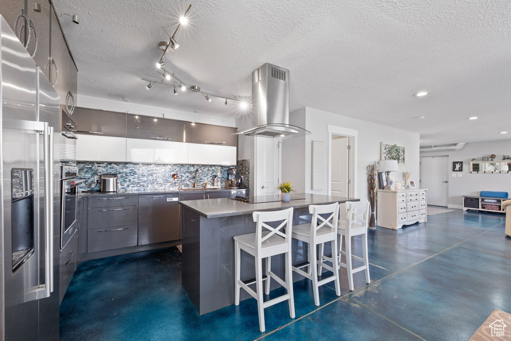 Kitchen featuring island range hood, appliances with stainless steel finishes, tasteful backsplash, modern cabinets, and a kitchen bar