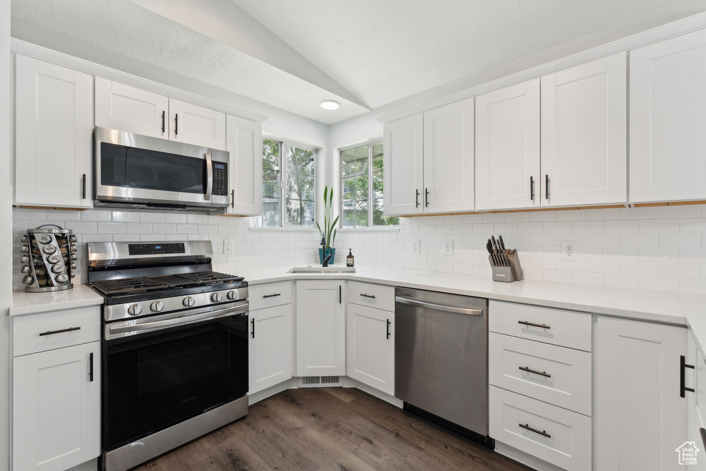 Kitchen featuring stainless steel appliances, lofted ceiling, white cabinetry, dark wood-type flooring, and backsplash