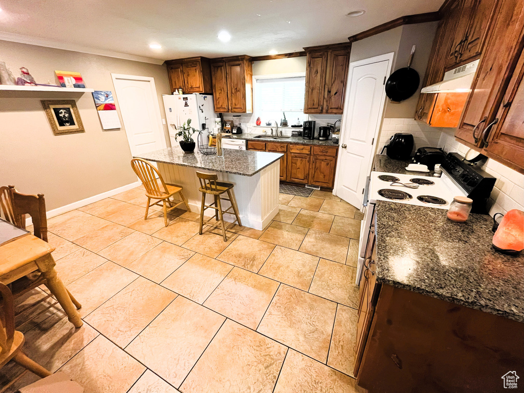 Kitchen with under cabinet range hood, white appliances, a sink, crown molding, and a breakfast bar area