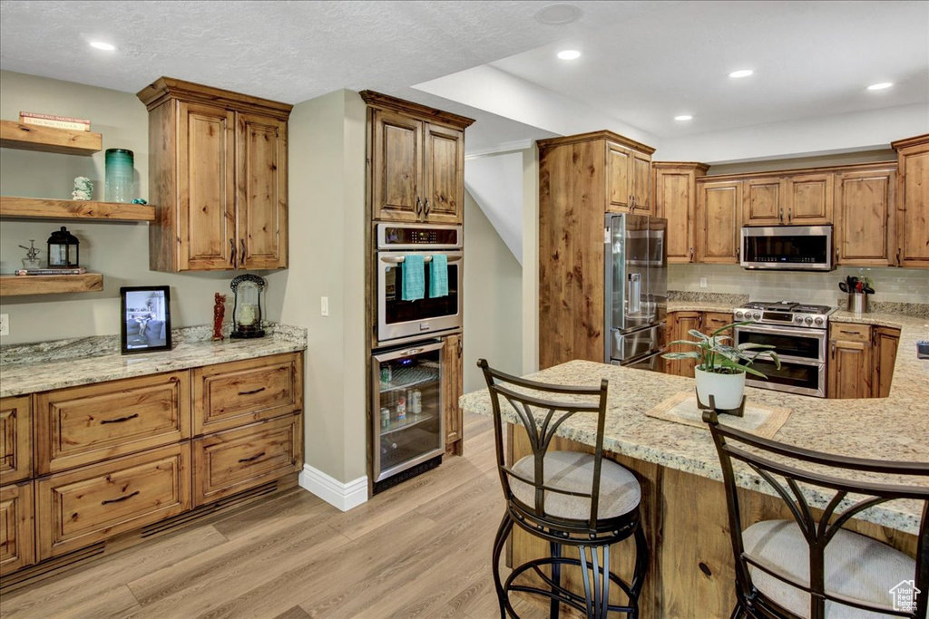 Kitchen with stainless steel appliances, beverage cooler, light wood-style flooring, light stone countertops, and a kitchen breakfast bar