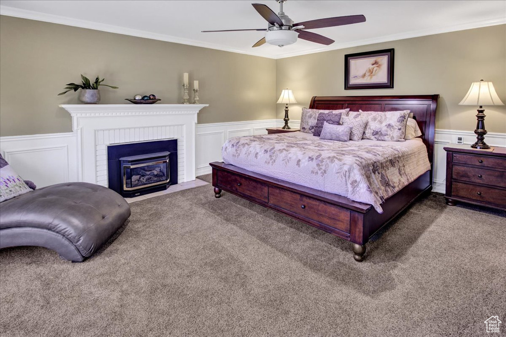 Carpeted bedroom with crown molding, wainscoting, a decorative wall, a brick fireplace, and a ceiling fan