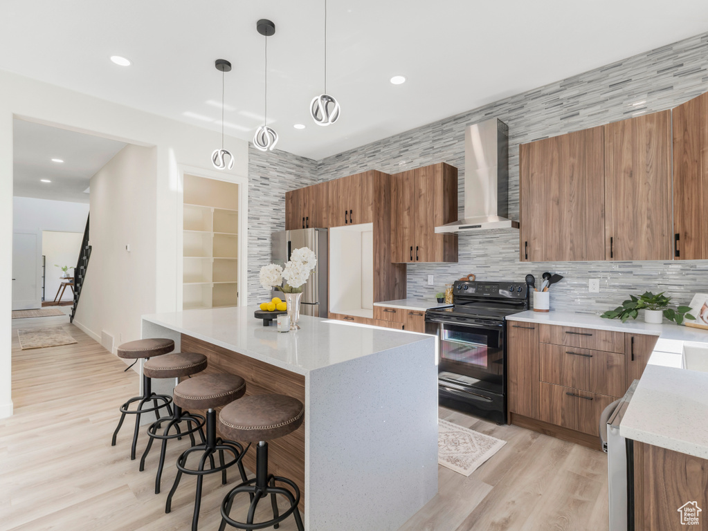Kitchen with black electric range oven, wall chimney exhaust hood, brown cabinets, a kitchen breakfast bar, and backsplash