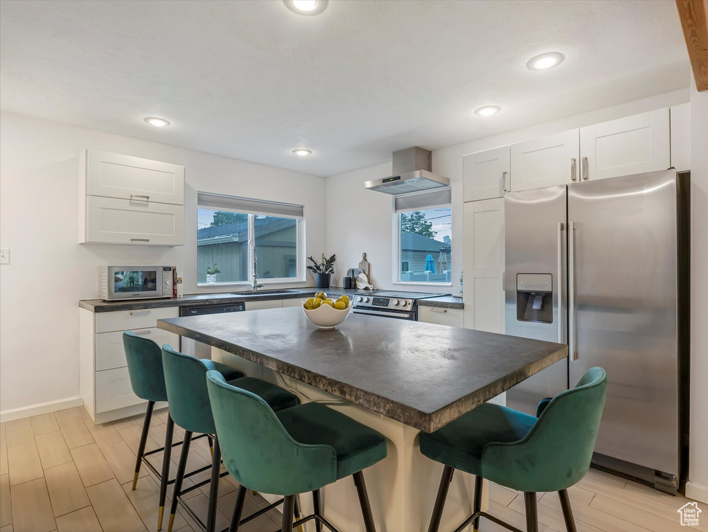 Kitchen featuring stainless steel refrigerator with ice dispenser, dark countertops, wall chimney exhaust hood, a kitchen bar, and white cabinetry