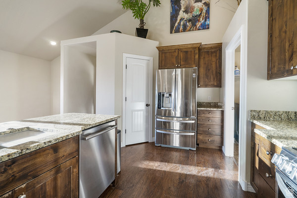 Kitchen with stainless steel appliances, dark wood-style flooring, light stone counters, vaulted ceiling, and dark brown cabinets