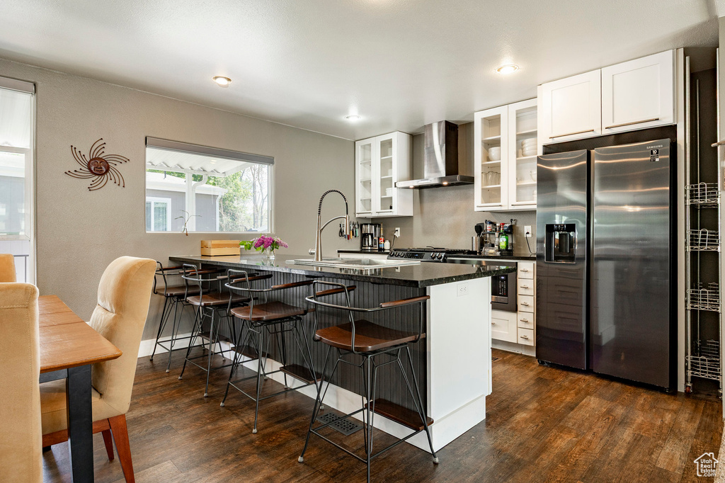 Kitchen featuring stainless steel fridge, wall chimney range hood, a kitchen breakfast bar, dark wood-type flooring, and dark countertops