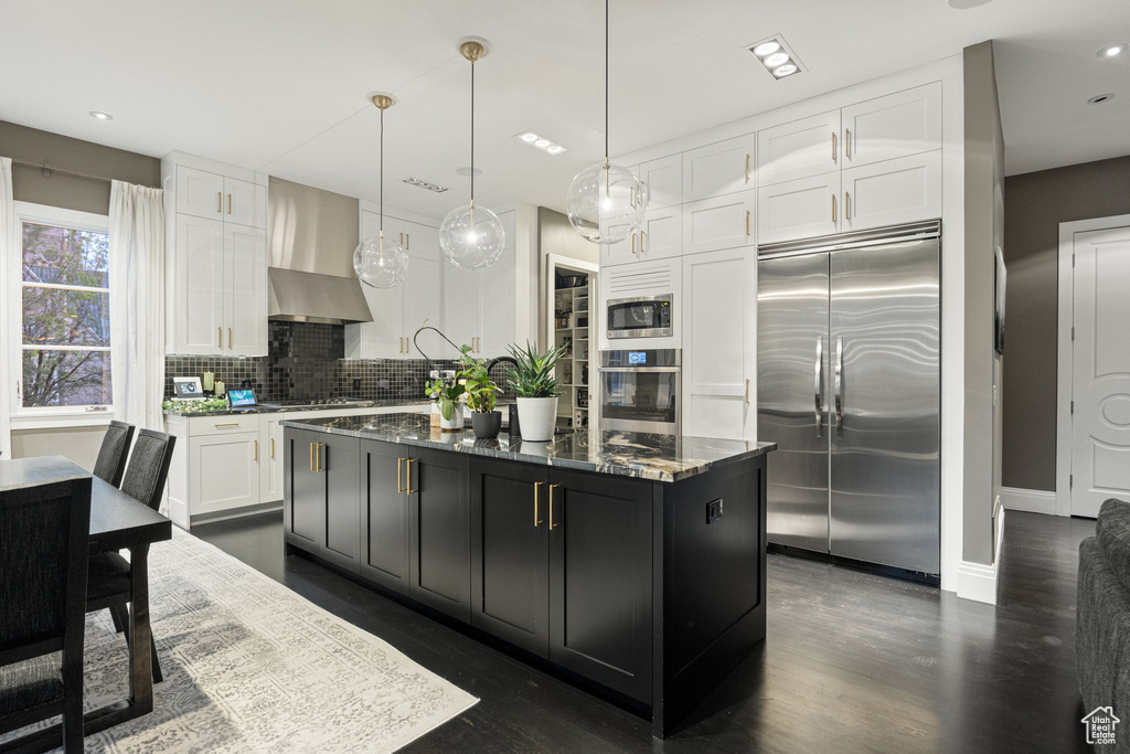 Kitchen featuring built in appliances, wall chimney exhaust hood, white cabinets, dark cabinets, and dark stone counters