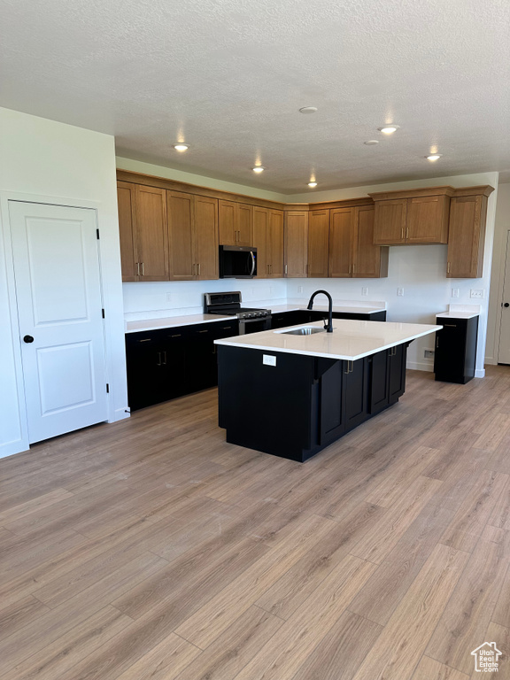 Kitchen with appliances with stainless steel finishes, a sink, brown cabinets, a kitchen island with sink, and light countertops