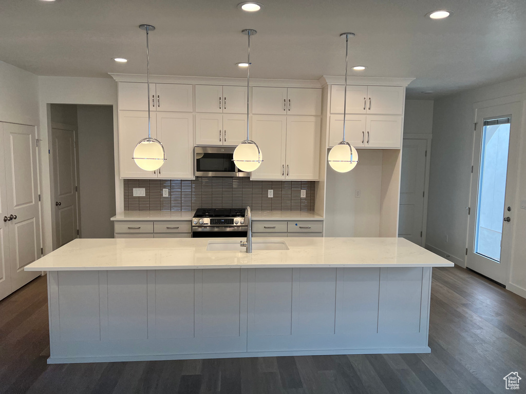 Kitchen with appliances with stainless steel finishes, dark wood-type flooring, backsplash, white cabinets, and a kitchen island with sink