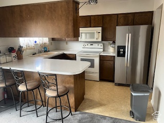 Kitchen with white appliances, a peninsula, light countertops, a breakfast bar, and dark brown cabinets