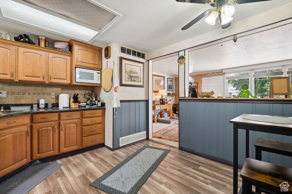 Kitchen with white microwave, light wood-type flooring, a ceiling fan, a wainscoted wall, and brown cabinetry