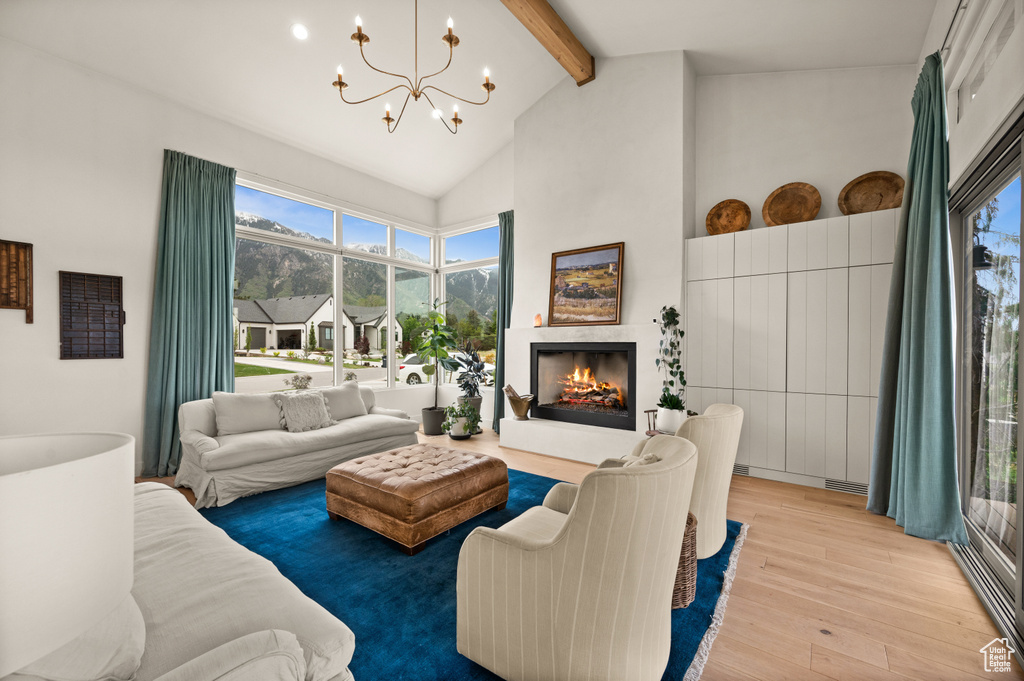 Living room featuring beam ceiling, light wood-style floors, a chandelier, high vaulted ceiling, and a warm lit fireplace