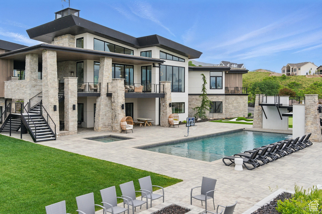 Back of property with an in-ground hot tub, a balcony, a patio area, a yard, and an outdoor pool