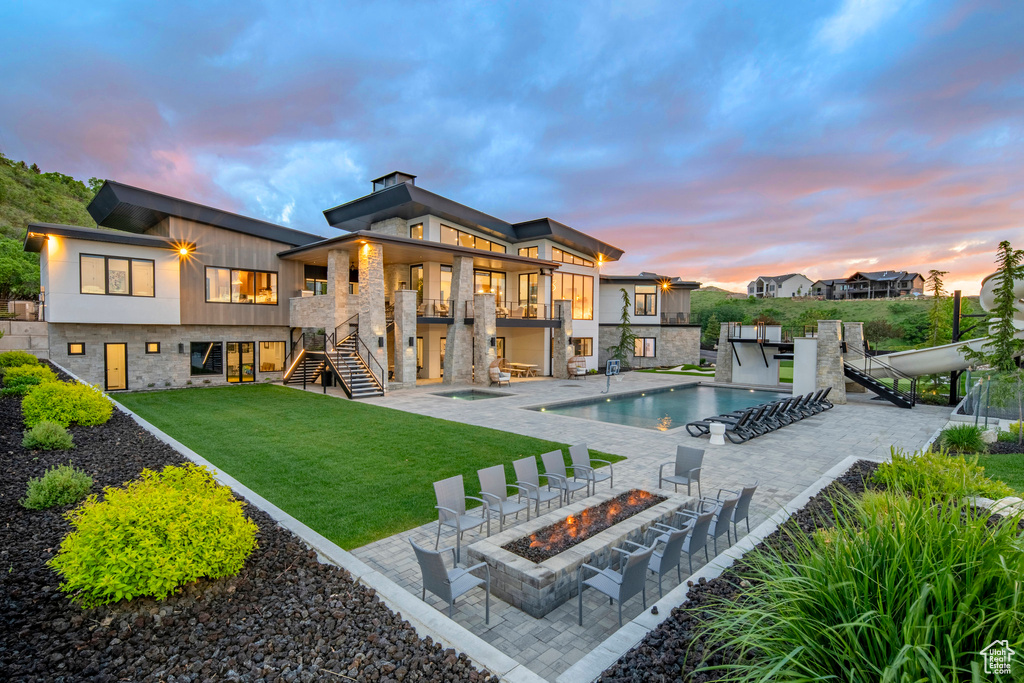 Back of house at dusk featuring stairs, a fire pit, a yard, a balcony, and an outdoor pool