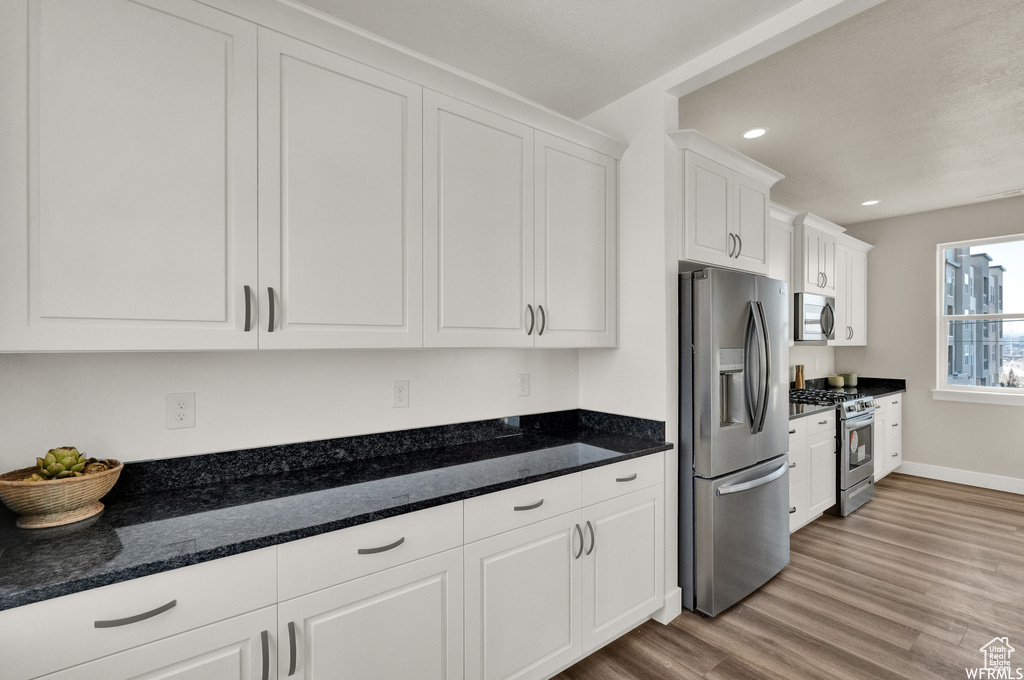 Kitchen with stainless steel appliances, white cabinetry, light wood-type flooring, recessed lighting, and dark stone counters