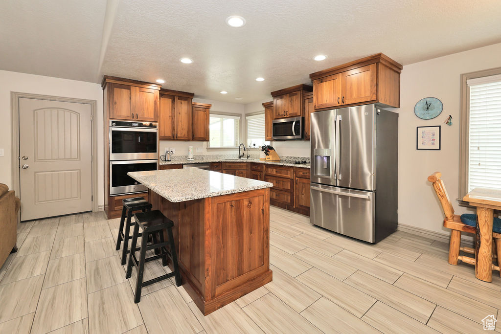 Kitchen with stainless steel appliances, a sink, a center island, light stone countertops, and brown cabinets