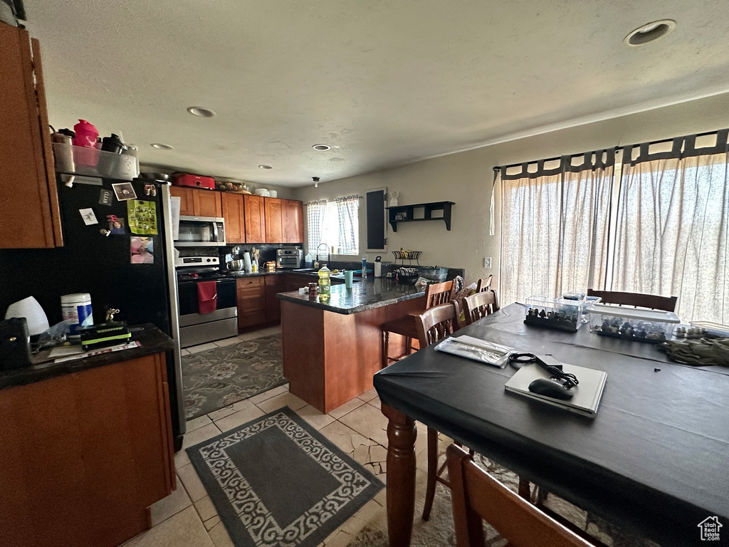 Kitchen featuring appliances with stainless steel finishes, a peninsula, dark countertops, brown cabinets, and light tile patterned flooring