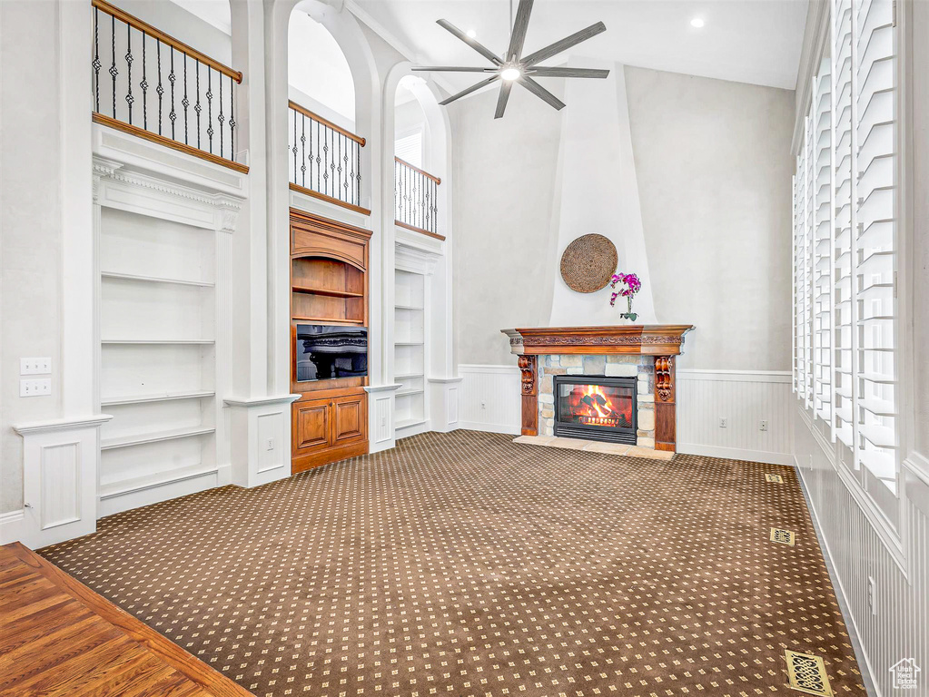 Unfurnished living room featuring a ceiling fan, built in shelves, carpet floors, a towering ceiling, and a fireplace with flush hearth