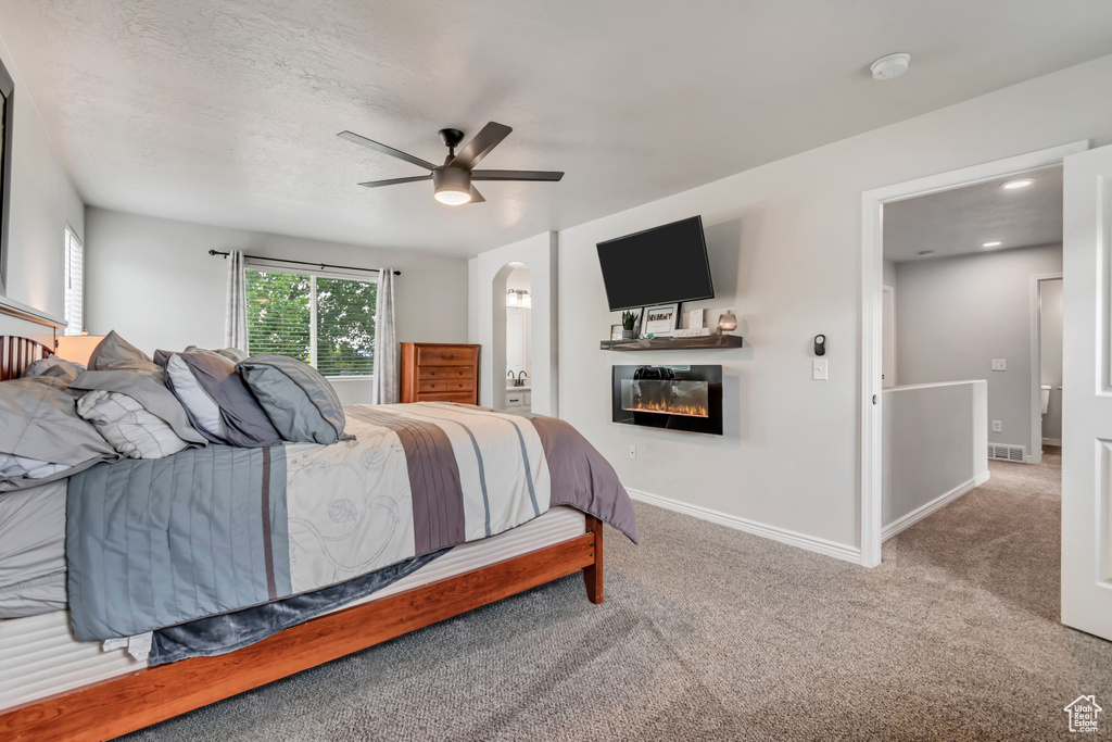 Carpeted bedroom featuring a glass covered fireplace, arched walkways, baseboards, ceiling fan, and ensuite bath