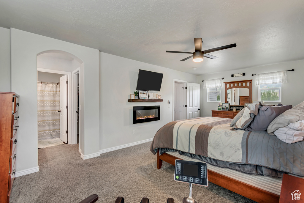 Carpeted bedroom with arched walkways, a glass covered fireplace, baseboards, ceiling fan, and ensuite bath