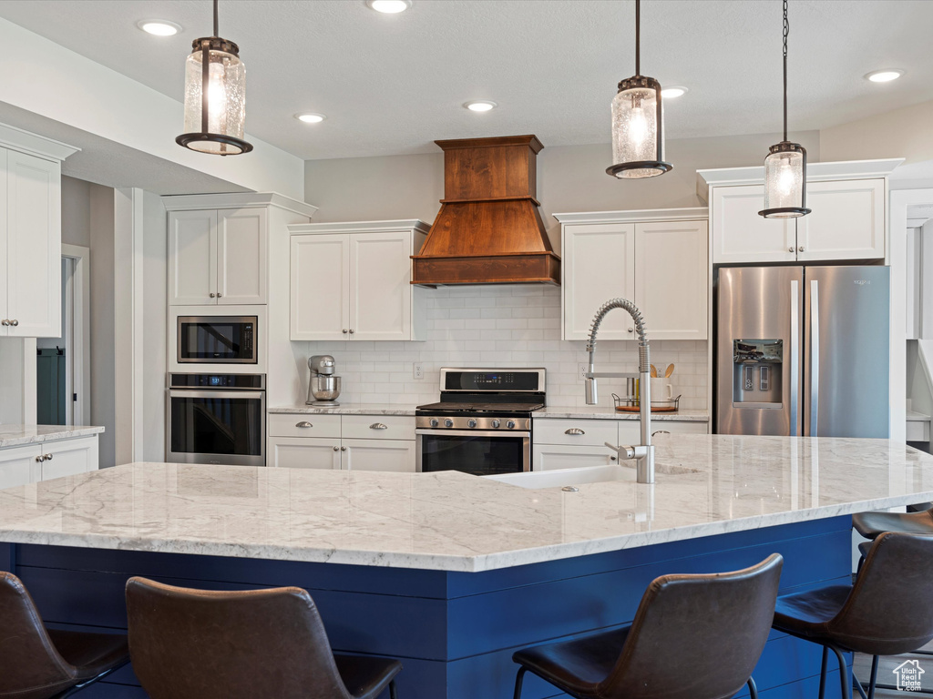 Kitchen with appliances with stainless steel finishes, premium range hood, white cabinets, tasteful backsplash, and a breakfast bar