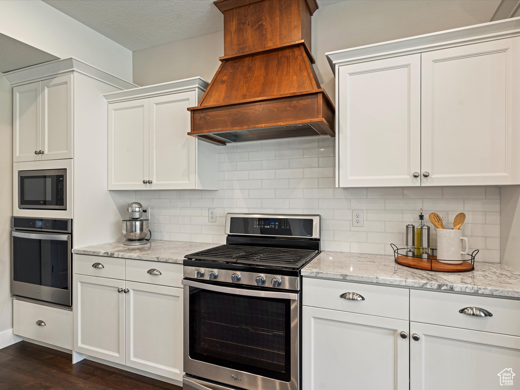 Kitchen with appliances with stainless steel finishes, custom range hood, white cabinetry, dark wood-style floors, and decorative backsplash
