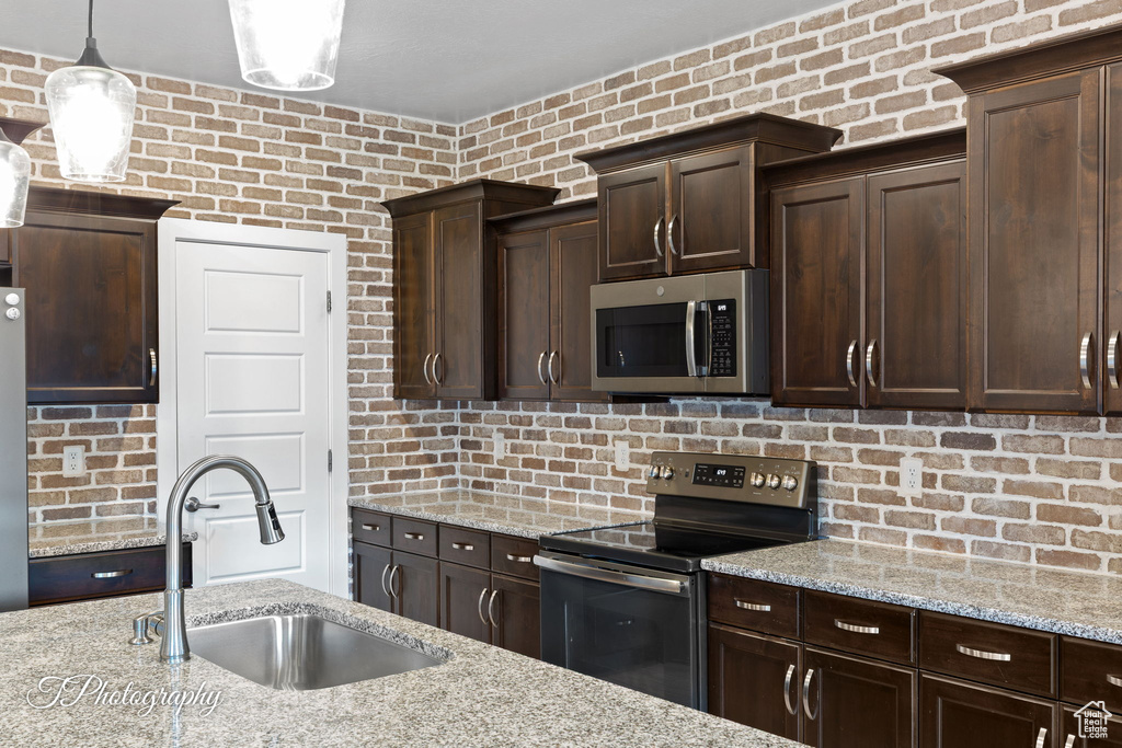 Kitchen with appliances with stainless steel finishes, a sink, dark brown cabinetry, light stone countertops, and brick wall