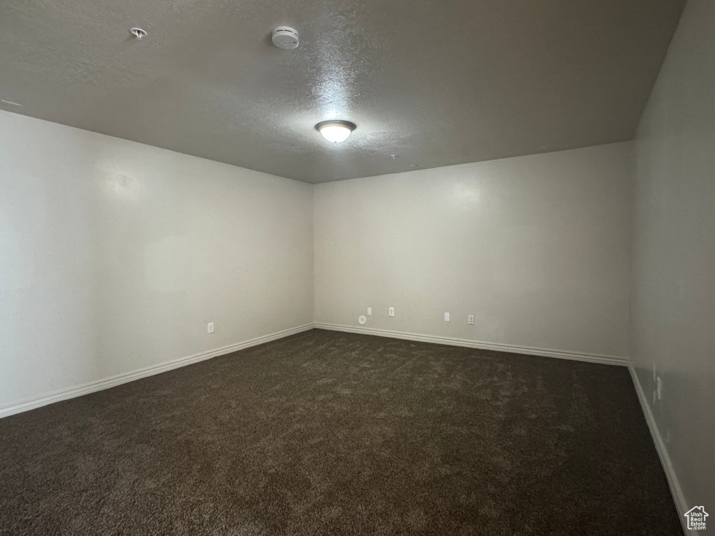 Empty room with a textured ceiling, dark carpet, baseboards, a smoke detector, and fire sprinkler in ceiling