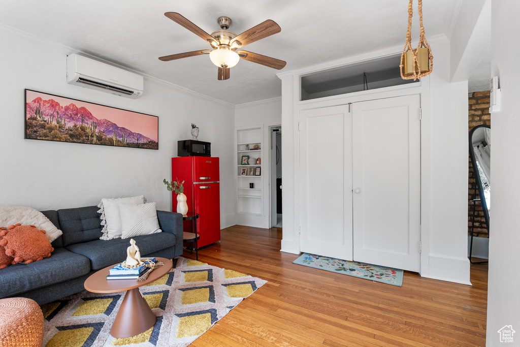 Living area with a wall mounted air conditioner, wood finished floors, ornamental molding, ceiling fan, and built in shelves