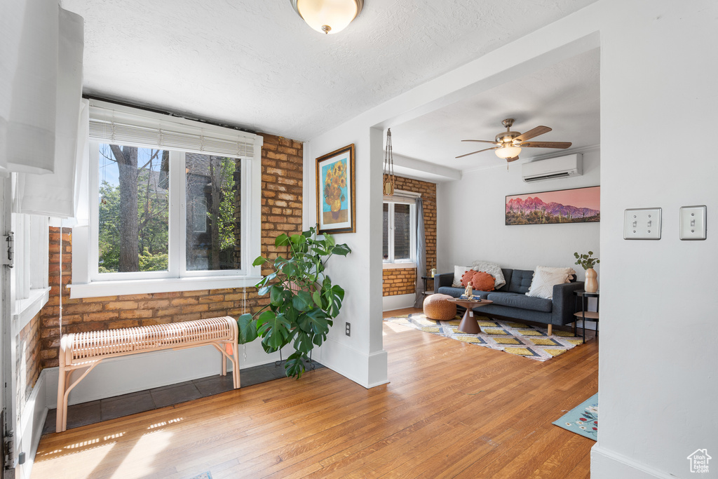 Living room with a wall unit AC, wood finished floors, ceiling fan, brick wall, and a textured ceiling