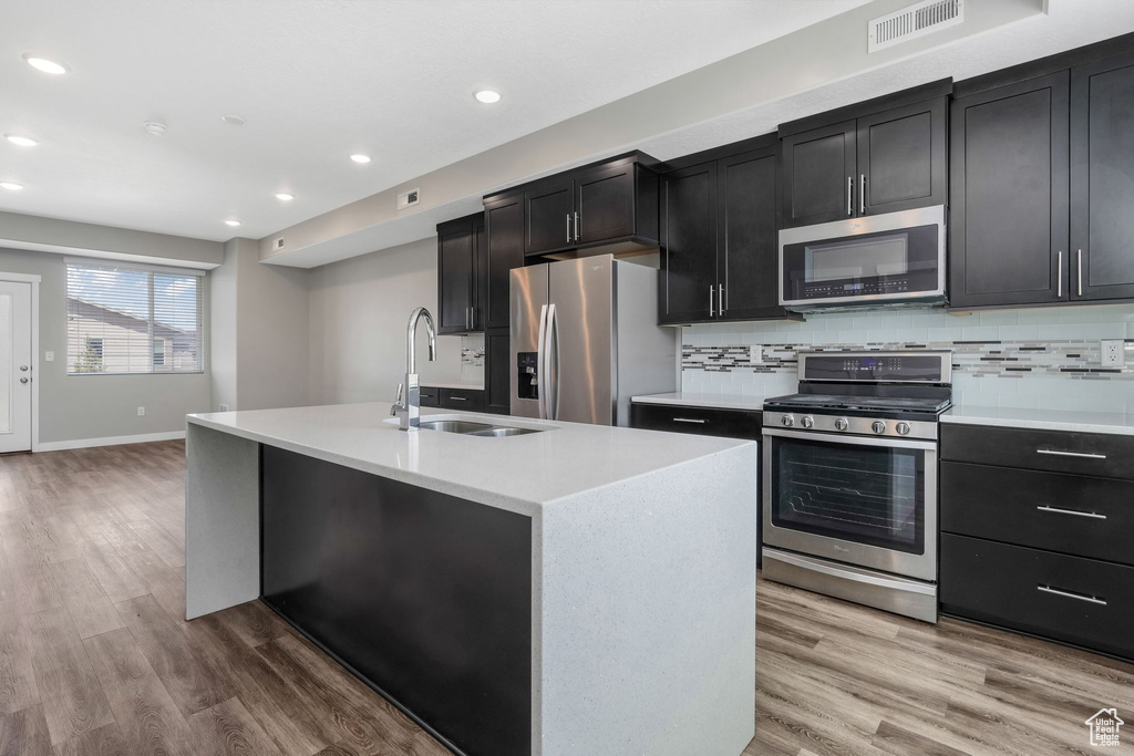 Kitchen with appliances with stainless steel finishes, a sink, dark cabinets, recessed lighting, and an island with sink