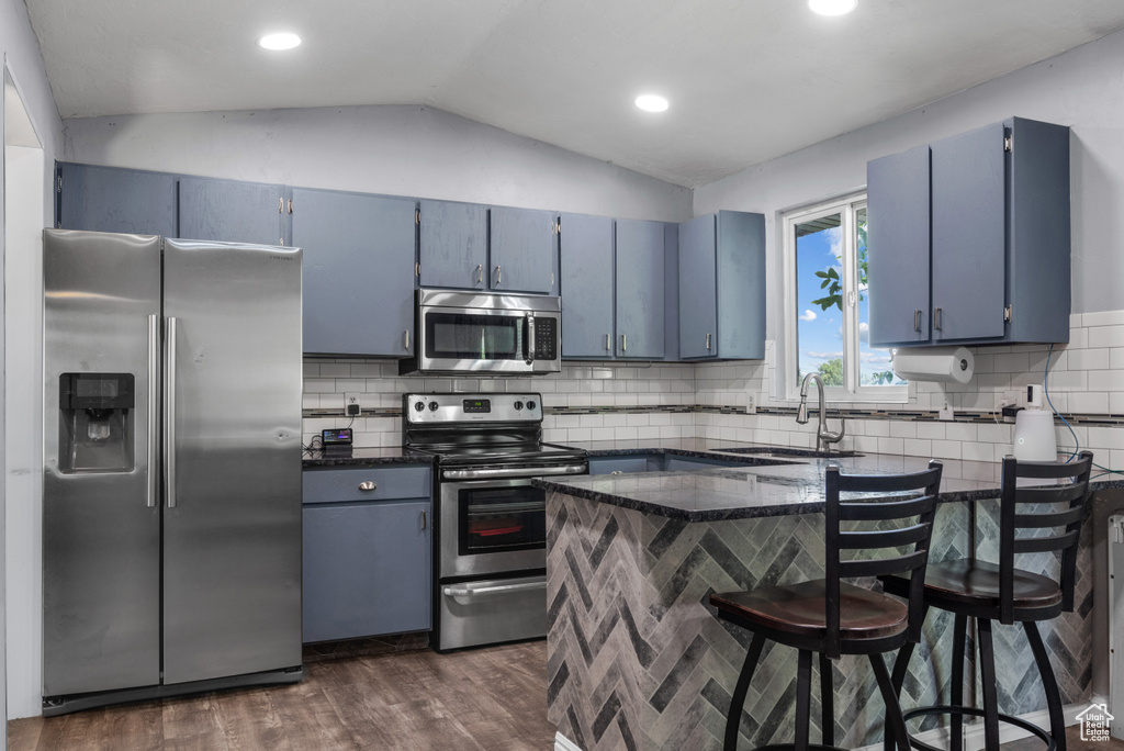 Kitchen with appliances with stainless steel finishes, a sink, blue cabinets, vaulted ceiling, and dark wood-style flooring