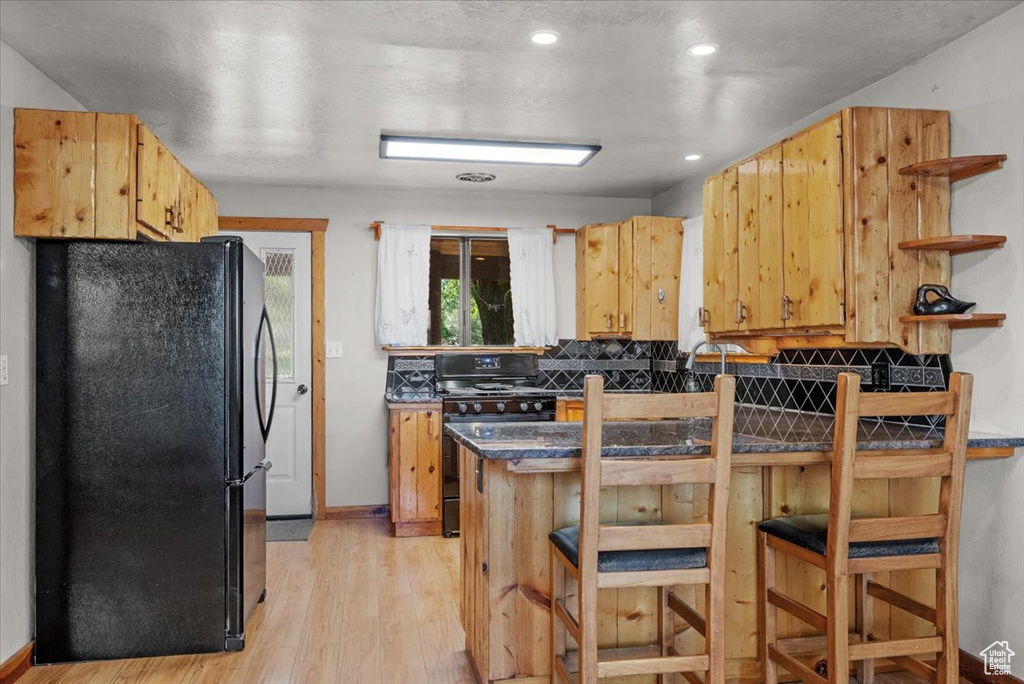 Kitchen with black appliances, open shelves, dark countertops, light wood finished floors, and a kitchen breakfast bar