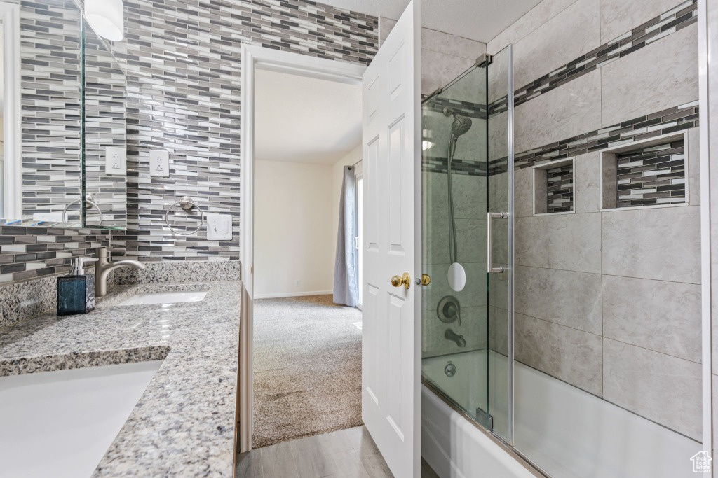 Full bath with bath / shower combo with glass door, baseboards, double vanity, decorative backsplash, and wood finished floors
