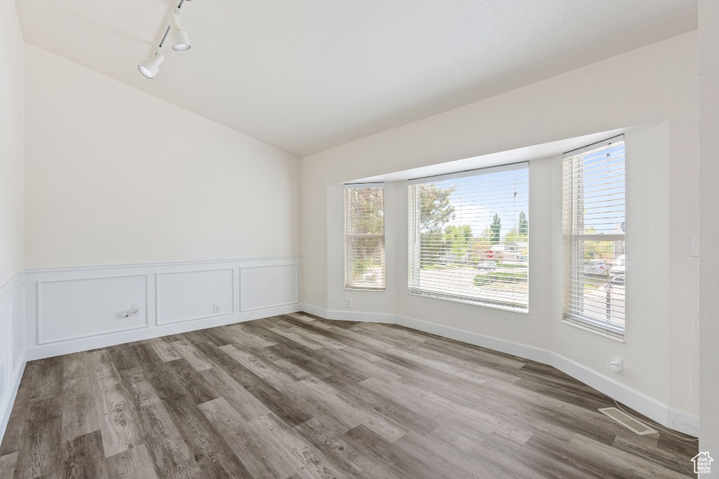 Empty room with rail lighting, lofted ceiling, wood finished floors, plenty of natural light, and a wainscoted wall