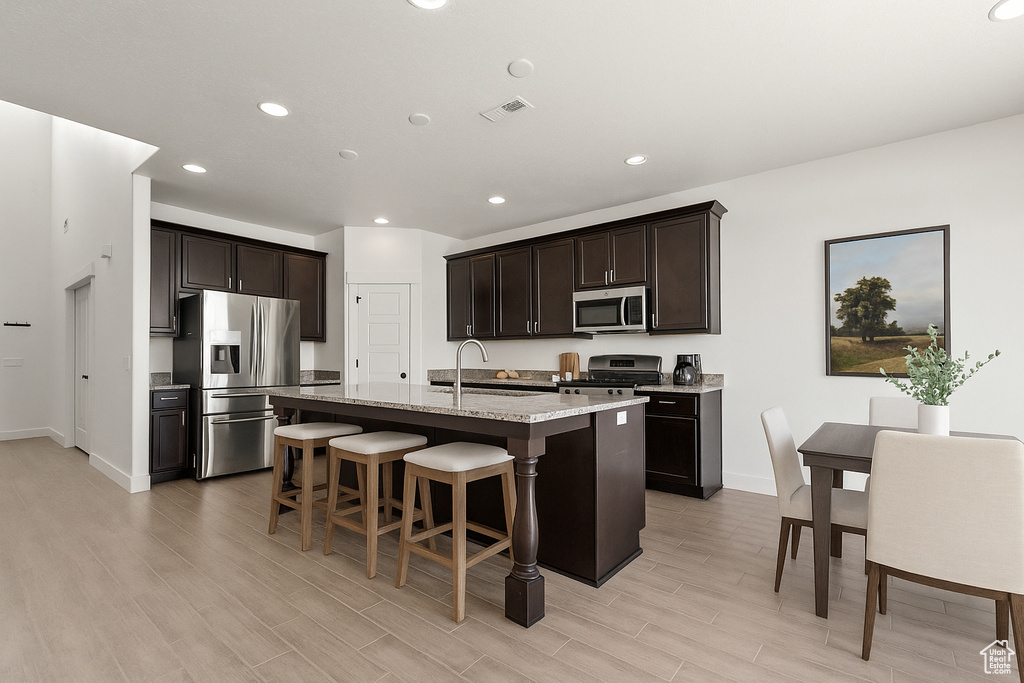 Kitchen featuring stainless steel appliances, light wood-type flooring, a kitchen island with sink, a breakfast bar, and dark brown cabinetry