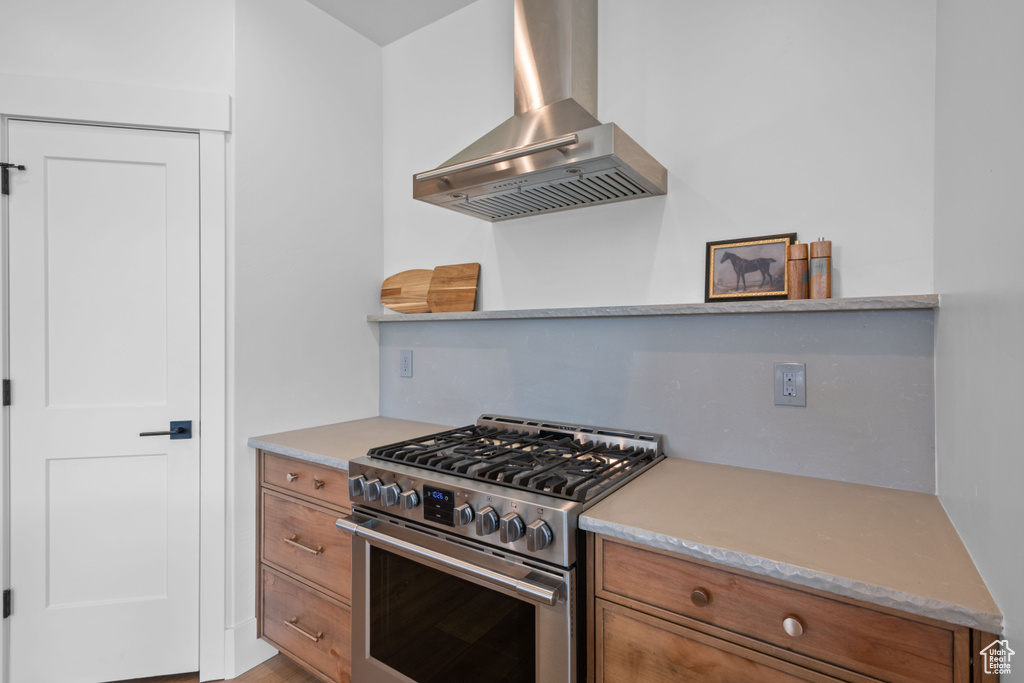 Kitchen with stainless steel stove, wall chimney range hood, brown cabinetry, and light countertops